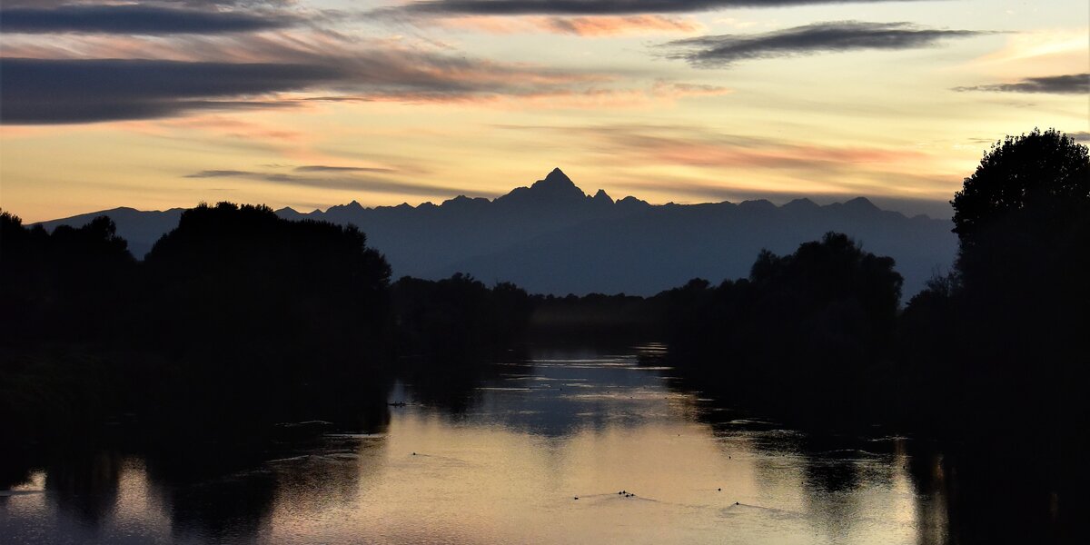 Veduta del Monviso al tramonto, con il profilo della montagna e delle cime alpine circostanti che si staglia contro un cielo dalle sfumature gialle, arancioni e rosate, attraversato da lunghe nuvole grigie. In primo piano scorre il fiume Po, che riflette i colori del cielo ed è fiancheggiato da alberi e vegetazione in controluce. Sulla superficie dell’acqua si distinguono alcuni uccelli acquatici.