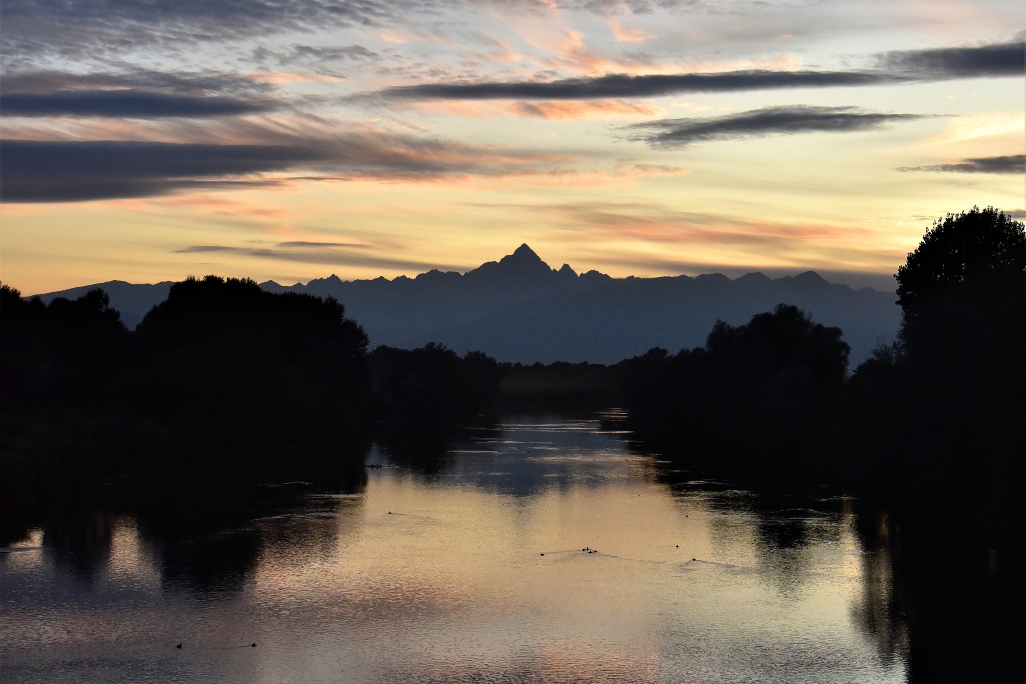 Veduta del Monviso al tramonto, con il profilo della montagna e delle cime alpine circostanti che si staglia contro un cielo dalle sfumature gialle, arancioni e rosate, attraversato da lunghe nuvole grigie. In primo piano scorre il fiume Po, che riflette i colori del cielo ed è fiancheggiato da alberi e vegetazione in controluce. Sulla superficie dell’acqua si distinguono alcuni uccelli acquatici.