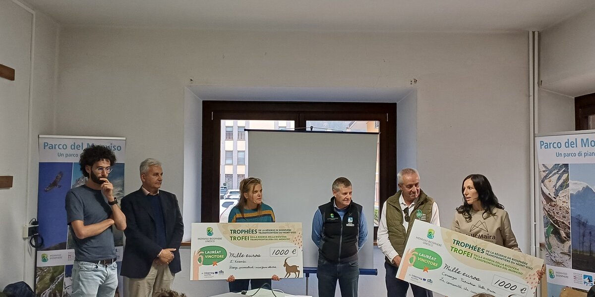 Cerimonia di premiazione dei Trofei della Riserva della Biosfera transfrontaliera del Monviso MaB UNESCO. In una sala conferenze, sei persone sono in piedi davanti al pubblico, tra pannelli informativi del Parco del Monviso. Due donne mostrano grandi assegni simbolici da mille euro, mentre gli altri partecipanti assistono alla premiazione. Al centro della sala si trovano un tavolo con un videoproiettore e uno schermo per le presentazioni.