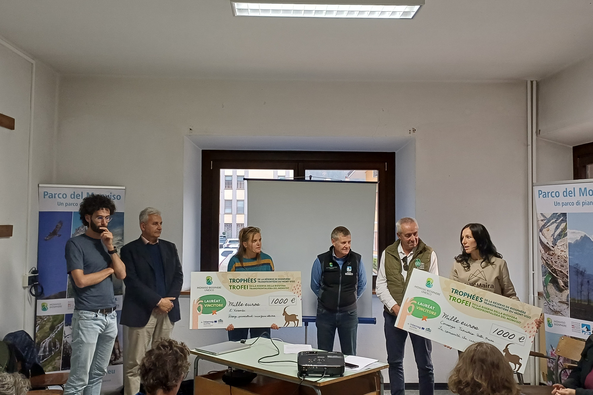 Cerimonia di premiazione dei Trofei della Riserva della Biosfera transfrontaliera del Monviso MaB UNESCO. In una sala conferenze, sei persone sono in piedi davanti al pubblico, tra pannelli informativi del Parco del Monviso. Due donne mostrano grandi assegni simbolici da mille euro, mentre gli altri partecipanti assistono alla premiazione. Al centro della sala si trovano un tavolo con un videoproiettore e uno schermo per le presentazioni.