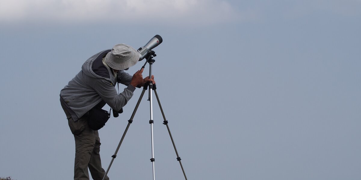 Un uomo in cima a Bric Lumbatera, con un cappello in testa, osserva il cielo con un cannocchiale poggiato su un treppiede.