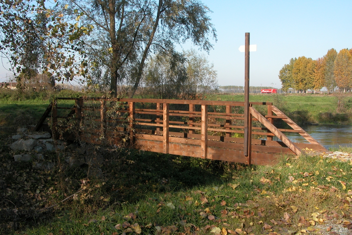 un ponte in legno con ringhiera sopra un corso d'acqua, intorno prati e alberi