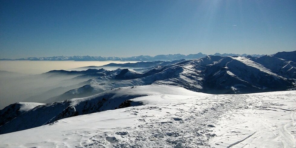 Dalla cima di una montagna panorama delle montagne circostanti completamente innevate al di sopra di una distesa di nuvole sulla sinistra. Cielo azzurro
