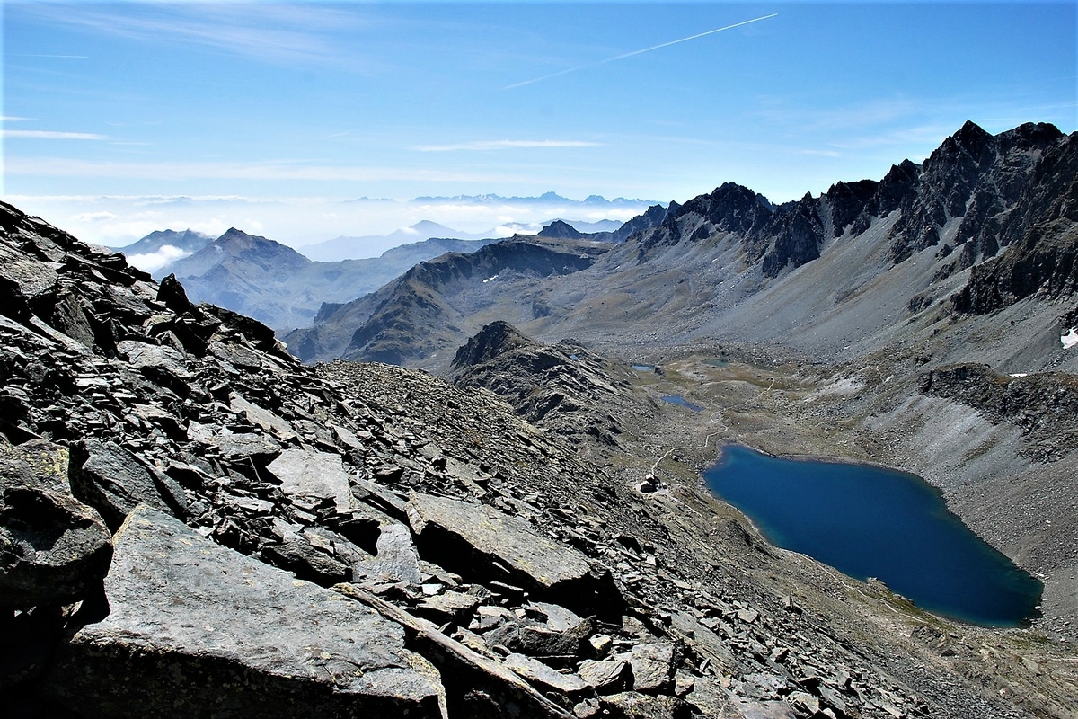 Immagine scattata da una parete montuosa, visibile in parte a sinistra: sulla destra un grande lago con a fianco una costruzione e sullo sfondo altre montagne.